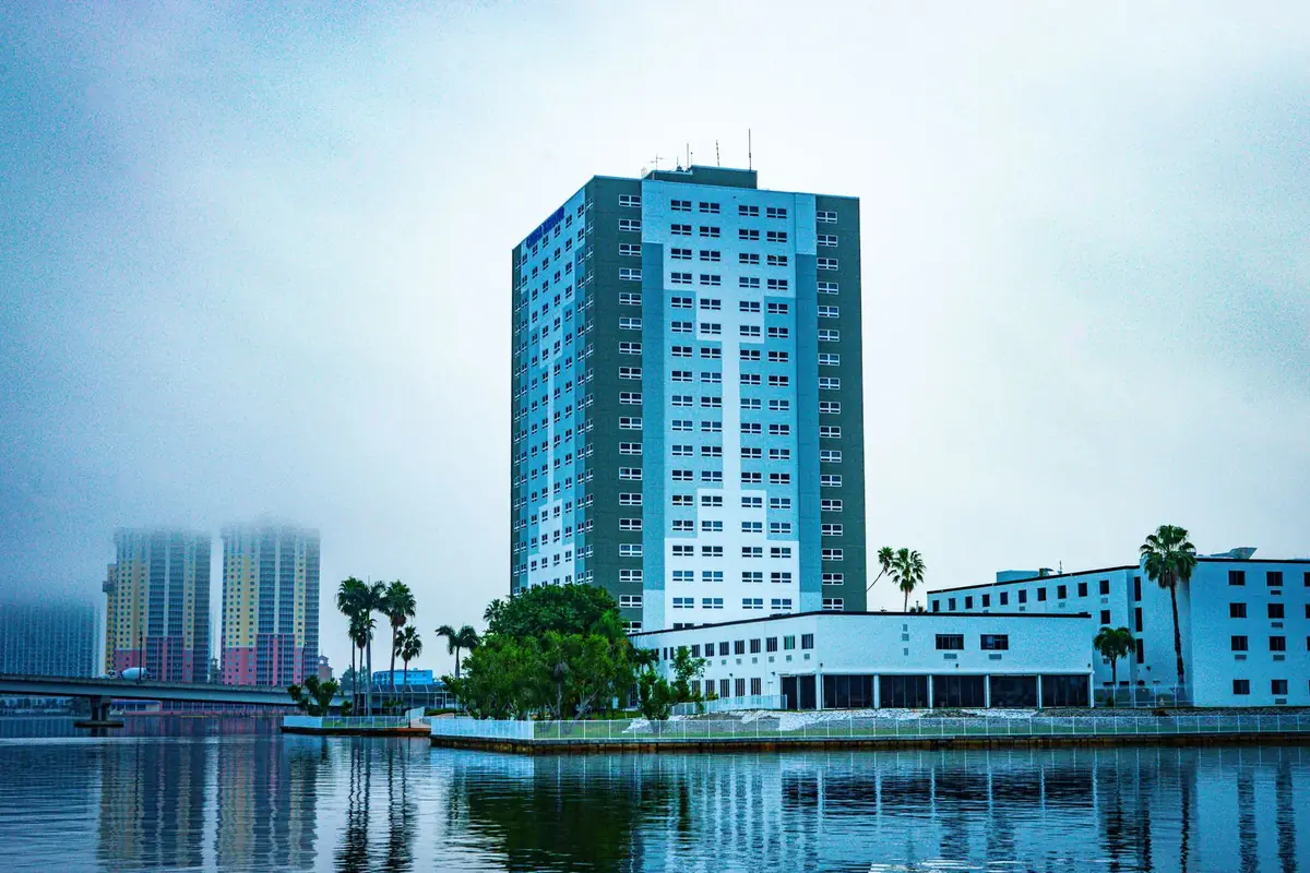 A high-rise building and palm trees on a riverfront, reflected in calm water