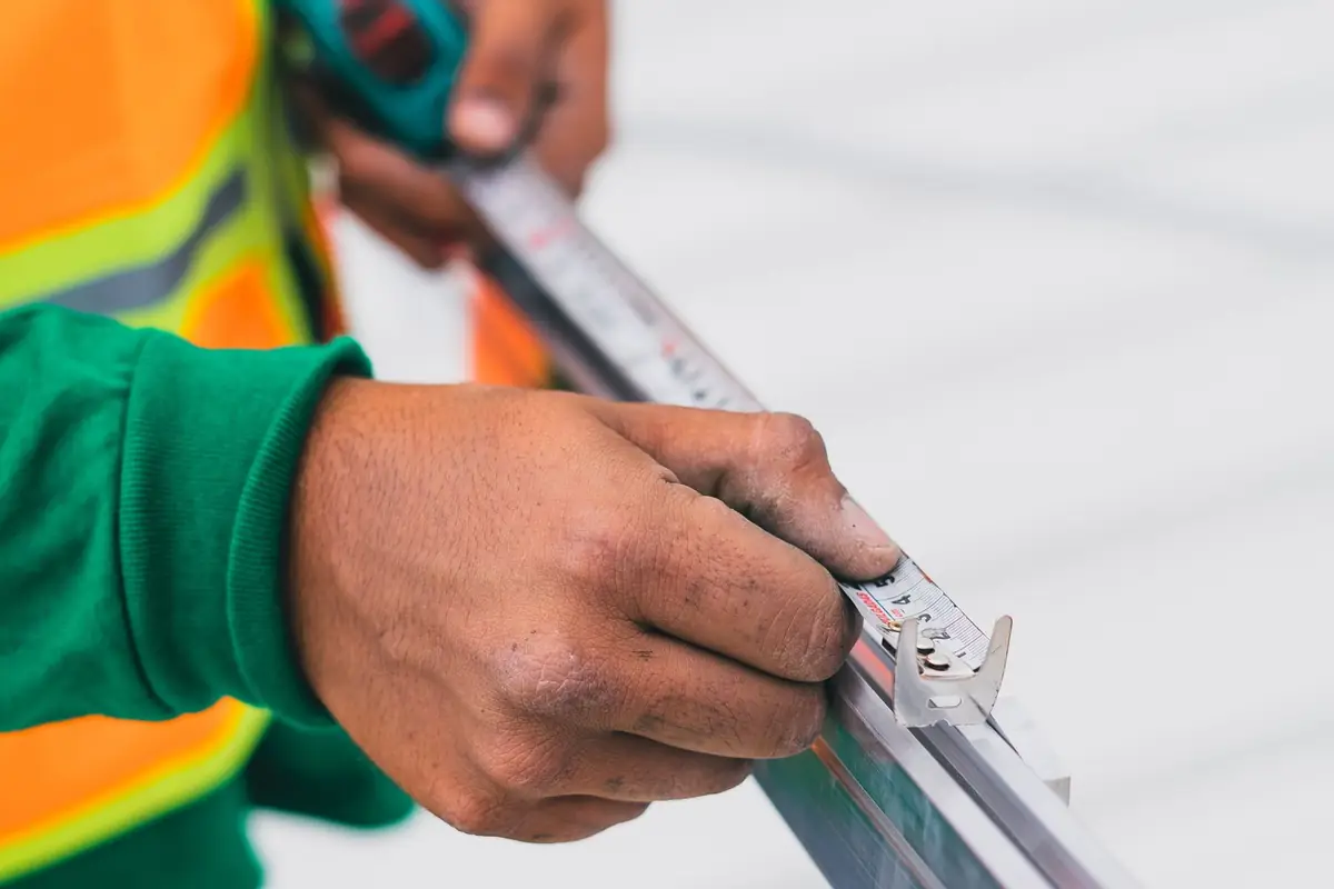 A worker in a hi-vis vest holding a tape measure against an aluminum frame profile
