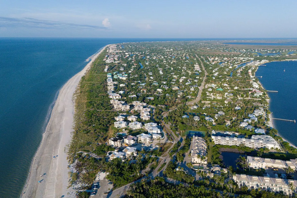 Aerial view of a sunny barrier island with homes among palms between a long sandy beach and a calm bay