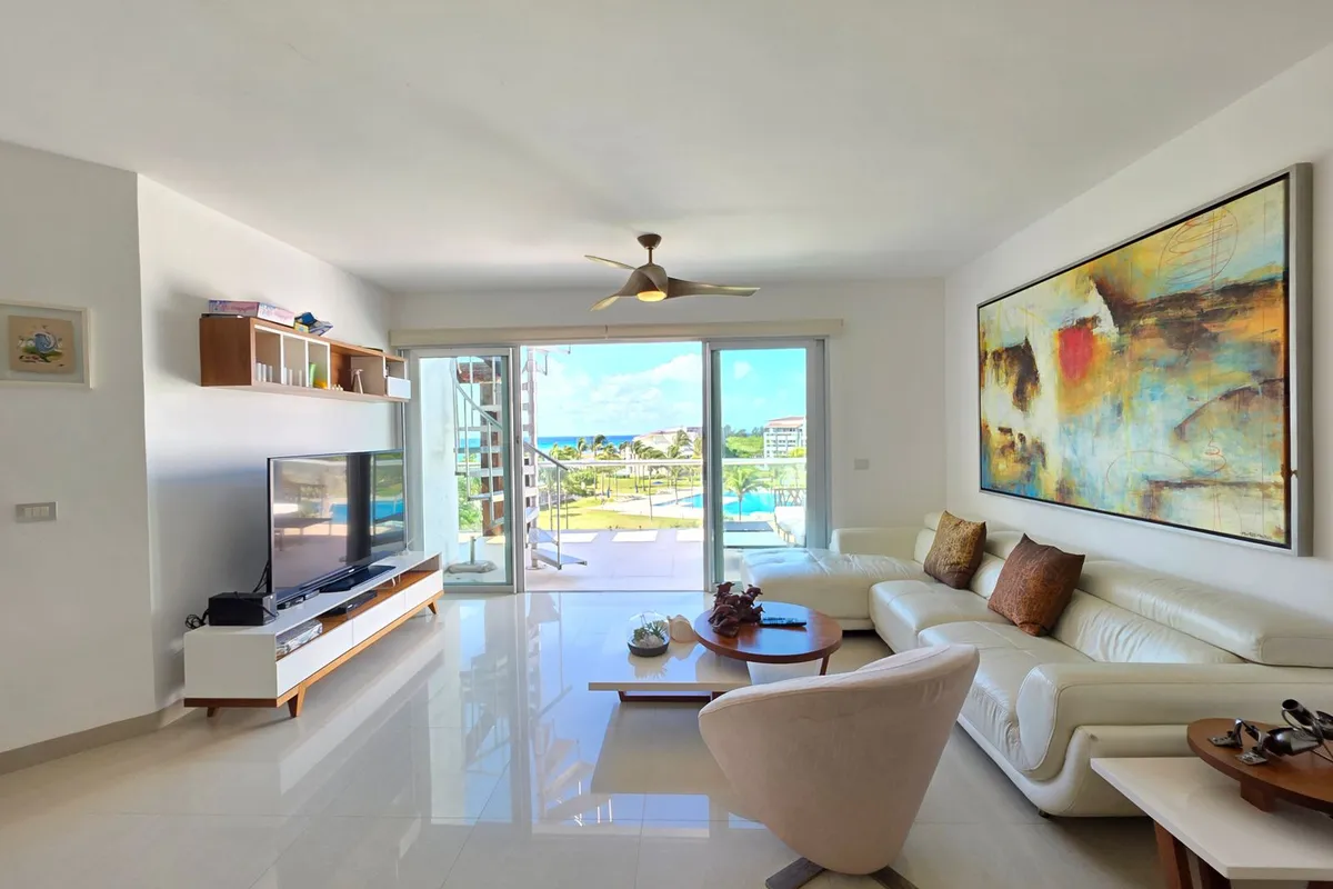 A bright, tiled living room with sliding glass doors open to a balcony overlooking a pool, palms and the sea